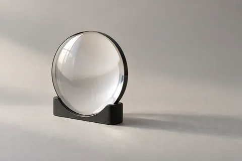 Una sola lente de vidrio óptico, de pie en un soporte negro sobre papel gris claro.